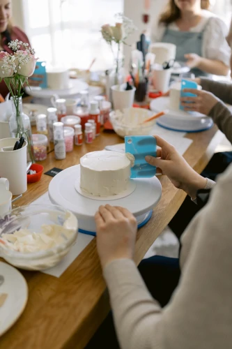 Hands decorating a cake at a shared workshop table with tools and icing